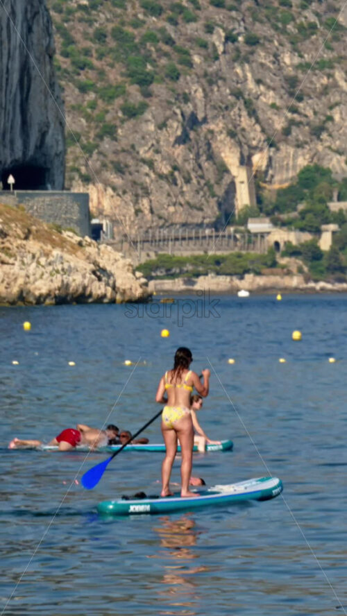 Video - Beaulieu-sur-Mer, France - July 5, 2024: Women paddleboarding on the sea with the beach on the background. Vertical