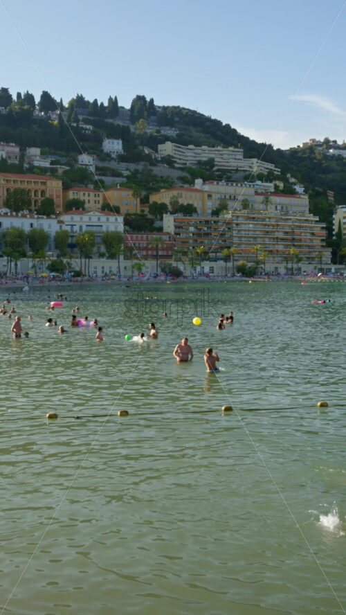 Video - Menton, France - September 4, 2024: People swimming in the sea with a view of the colourful buildings in the city. Vertical