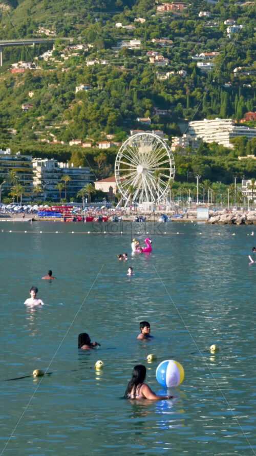 Video - Menton, France - September 4, 2024: People swimming in the sea with a view of the colourful buildings in the city. Vertical
