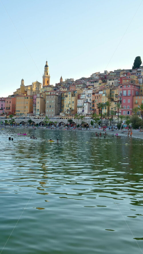 Video - Menton, France - September 4, 2024: People swimming in the sea with a view of the colourful buildings in the city. Vertical