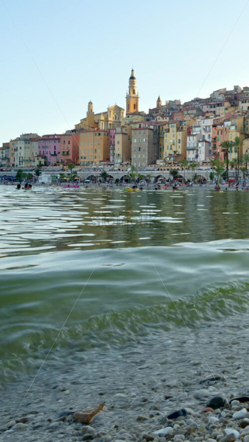 Video - Menton, France - September 4, 2024: People swimming in the sea with a view of the colourful buildings in the city. Vertical