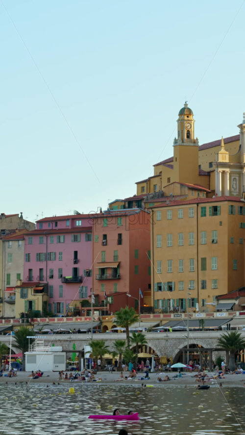 Video - Menton, France - September 4, 2024: People swimming in the sea with a view of the colourful buildings in the city. Vertical