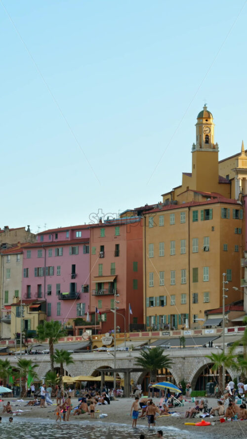 Video - Menton, France - September 4, 2024: People swimming in the sea with a view of the colourful buildings in the city. Vertical