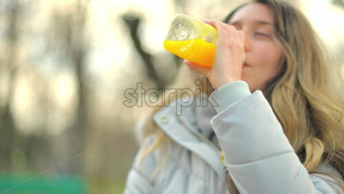 Video - Woman in a puffer jacket drinking orange juice in a park