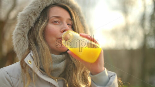 Video - Woman in a puffer jacket drinking orange juice in a park