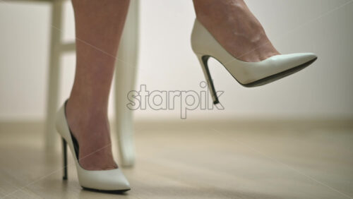 Video - Close up of the feet of a woman wearing white high heels sitting on a chair