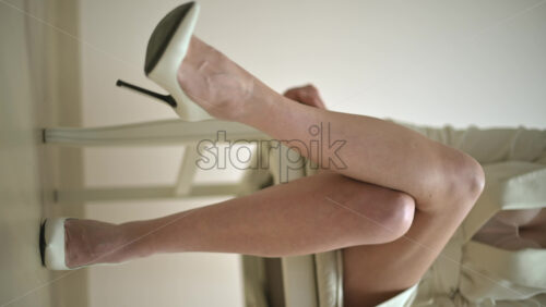 Video - Close up of the feet of a woman wearing white high heels sitting on a chair. Vertical