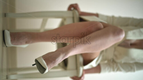 Video - Close up of the feet of a woman wearing white high heels sitting on a chair. Vertical