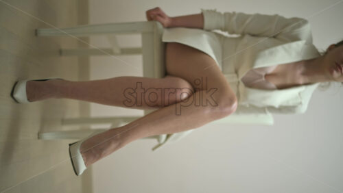Video - Woman in a white dress wearing white high heels sitting on a chair. Vertical