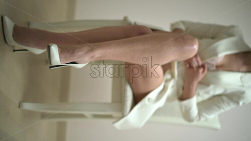 Video - Woman in a white dress wearing white high heels sitting on a chair. Vertical