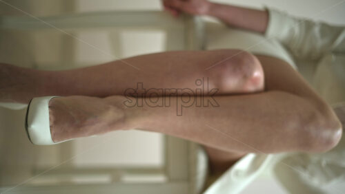 Video - Woman in a white dress wearing white high heels sitting on a chair. Vertical