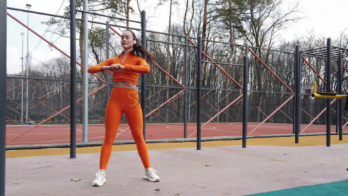 Video - Woman in orange sportswear warming up near a basketball court outside