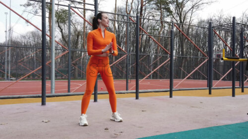 Video - Woman in orange sportswear warming up near a basketball court outside
