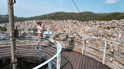 Video - A woman in white t-shirt on the view point is filming on smartphone, buildings, hills on the background, Kavala, Greece