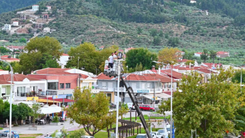Video - THASOS, GREECE - SEPTEMBER 23, 2020: Two men on a crane repairing a lamp post in the town, greenery and buildings on the background