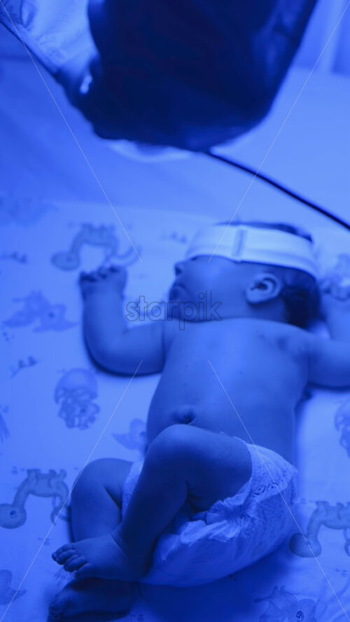 Video - Close up of a newborn baby moving around on a crib mattress, wearing a diaper with calming blue lighting. Vertical