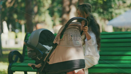 Video - Young mother sitting on a park bench next to a stroller, enjoying a peaceful outdoor moment with her baby