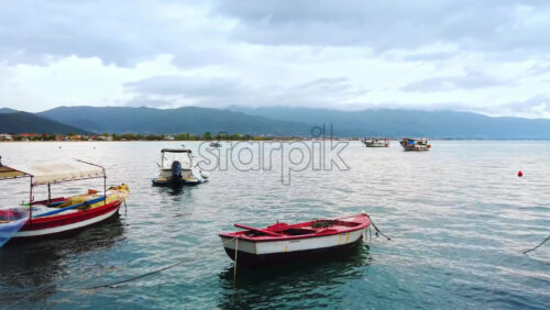 Video - Moored boats on the shore of the Aegean sea, town and hills on the background, cloudy sky in Stavros, Greece