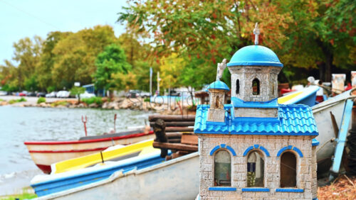 Video - Small model of a church with blue roof and cupolas on the beach of Stavros, greenery and boats on the background, Greece