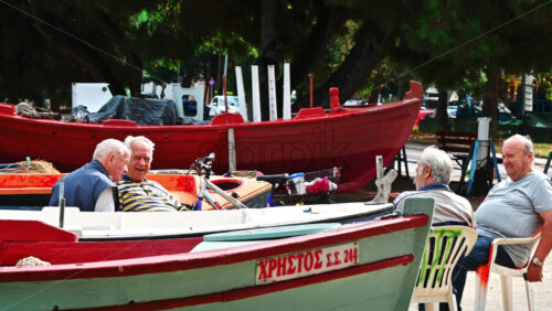 Video - STAVROS, GREECE - SEPTEMBER 26, 2020: Four old men resting on the beach with beached boats around, street on the background