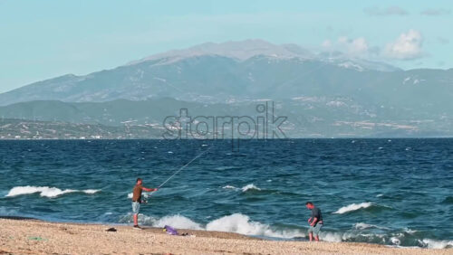 Video - OLYMPIADA, GREECE - SEPTEMBER 27, 2020: Beach of Aegean sea with two fishermen, waves with foam, land in the distance