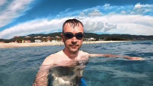 Video - Man in swimming goggles in the water of the Aegean sea holding camera, nature on the background. Slow motion. Greece