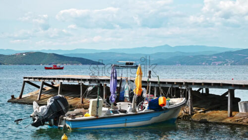 Video - Beached metal motorized colored boat on a pier on Aegean sea coast, hills and a town on the background in Ouranoupolis, Greece