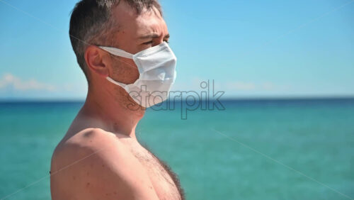 Video - Portrait of a man in white medical mask on the Aegean sea shore in Greece, water on the background