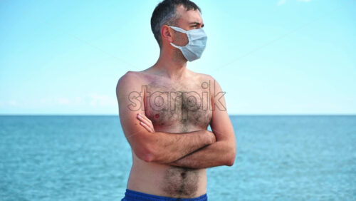 Video - Portrait of a man in white medical mask on the Aegean sea shore in Greece, water on the background