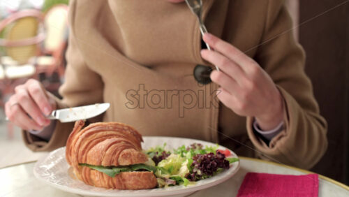 Video - Woman in brown coat eating a poached egg croissant with salad at a restaurant