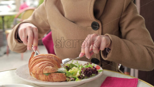 Video - Woman in brown coat eating a poached egg croissant with salad at a restaurant