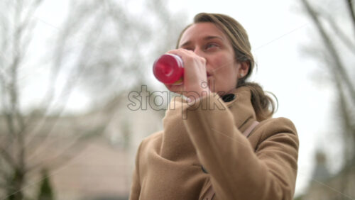 Video - Woman in brown coat drinking a pink beverage outside