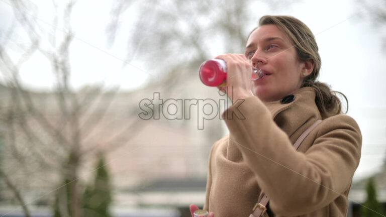 Video - Woman in brown coat drinking a pink beverage outside
