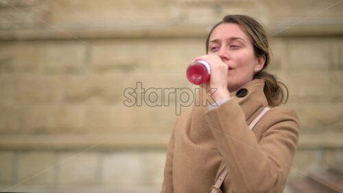 Video - Woman in brown coat drinking a pink beverage outside