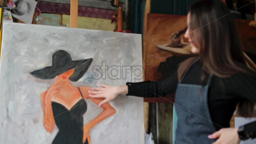 Video - Woman painter wearing black clothes painting woman model face with hat drawing with red brush at a workshop