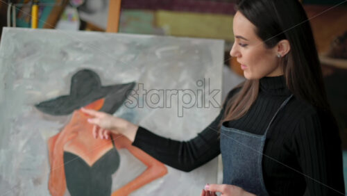 Video - Woman painter wearing black clothes painting woman model face with hat drawing with red brush at a workshop