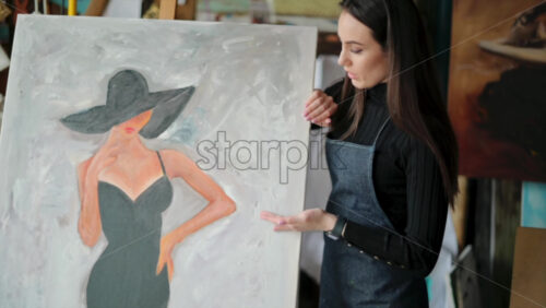 Video - Woman painter wearing black clothes painting woman model face with hat drawing with red brush at a workshop