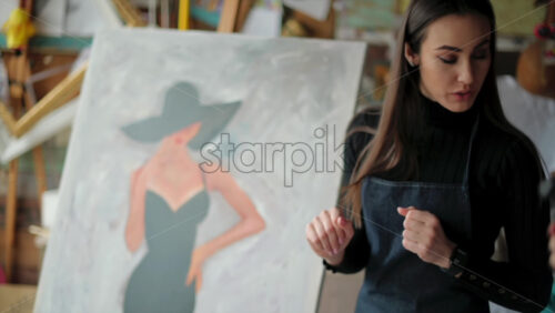 Video - Woman painter wearing black clothes painting woman model face with hat drawing with red brush at a workshop