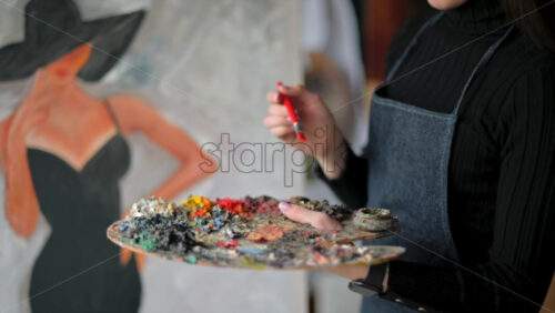 Video - Woman painter wearing black clothes painting woman model face with hat drawing with red brush at a workshop