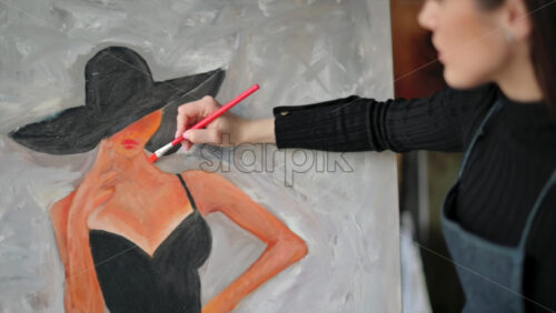 Video - Woman painter wearing black clothes painting woman model face with hat drawing with red brush at a workshop
