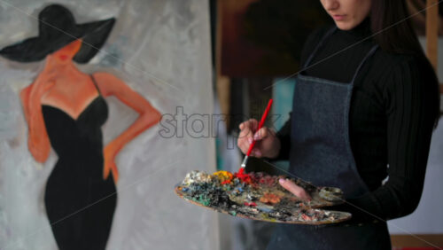 Video - Woman painter wearing black clothes painting woman model face with hat drawing with red brush at a workshop