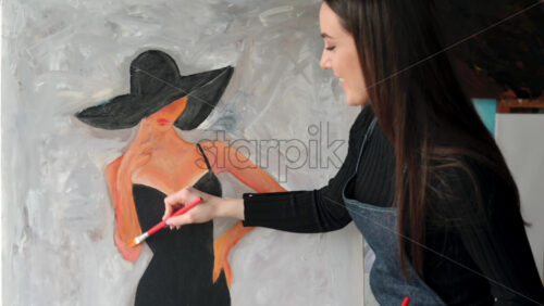 Video - Woman painter wearing black clothes painting woman model face with hat drawing with red brush at a workshop