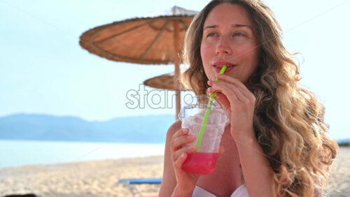 Video - Caucasian woman drinking a red cocktail on the beach. Sea, umbrellas. Slow motion