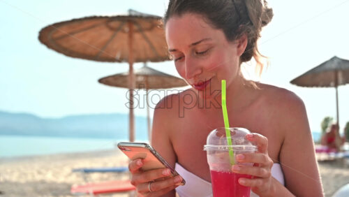Video - Caucasian woman drinking a red cocktail and is on her smartphone on the beach. Sea, umbrellas. Slow motion
