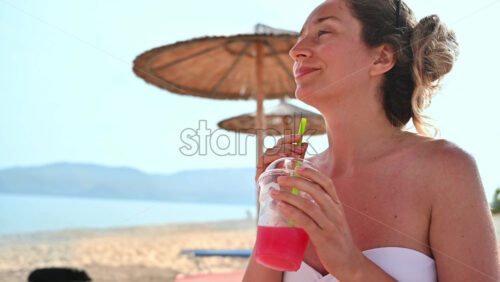 Video - Caucasian woman drinking a red cocktail on the beach. Sea, umbrellas. Slow motion