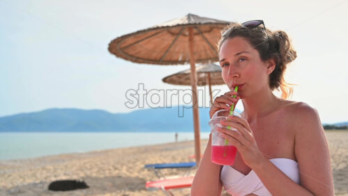 Video - Caucasian woman drinking a red cocktail on the beach. Sea, umbrellas. Slow motion