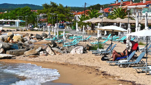 Video - NIKITI, GREECE - OCTOBER 8, 2020: Beach on Aegean sea coast with resting people, umbrellas and sunbeds, rocks in the water, embankment street