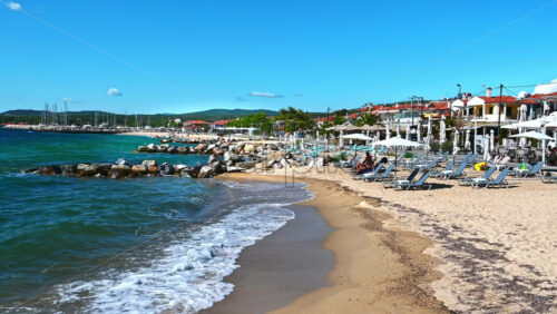 Video - NIKITI, GREECE - OCTOBER 8, 2020: Beach on Aegean sea coast with resting people, umbrellas and sunbeds, rocks in the water, embankment street
