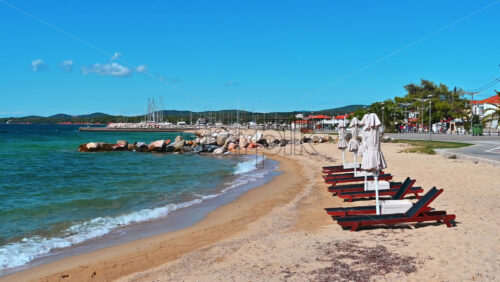 Video - Beach on Aegean sea coast with closed umbrellas and sunbeds, rocks in the water, embankment street, sea port and green hills in the distance in Nikiti, Greece