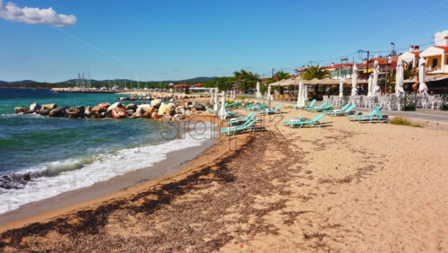 Video - Beach on Aegean sea coast with closed umbrellas and sunbeds, rocks in the water, embankment street, sea port and green hills in the distance in Nikiti, Greece. Slow motion